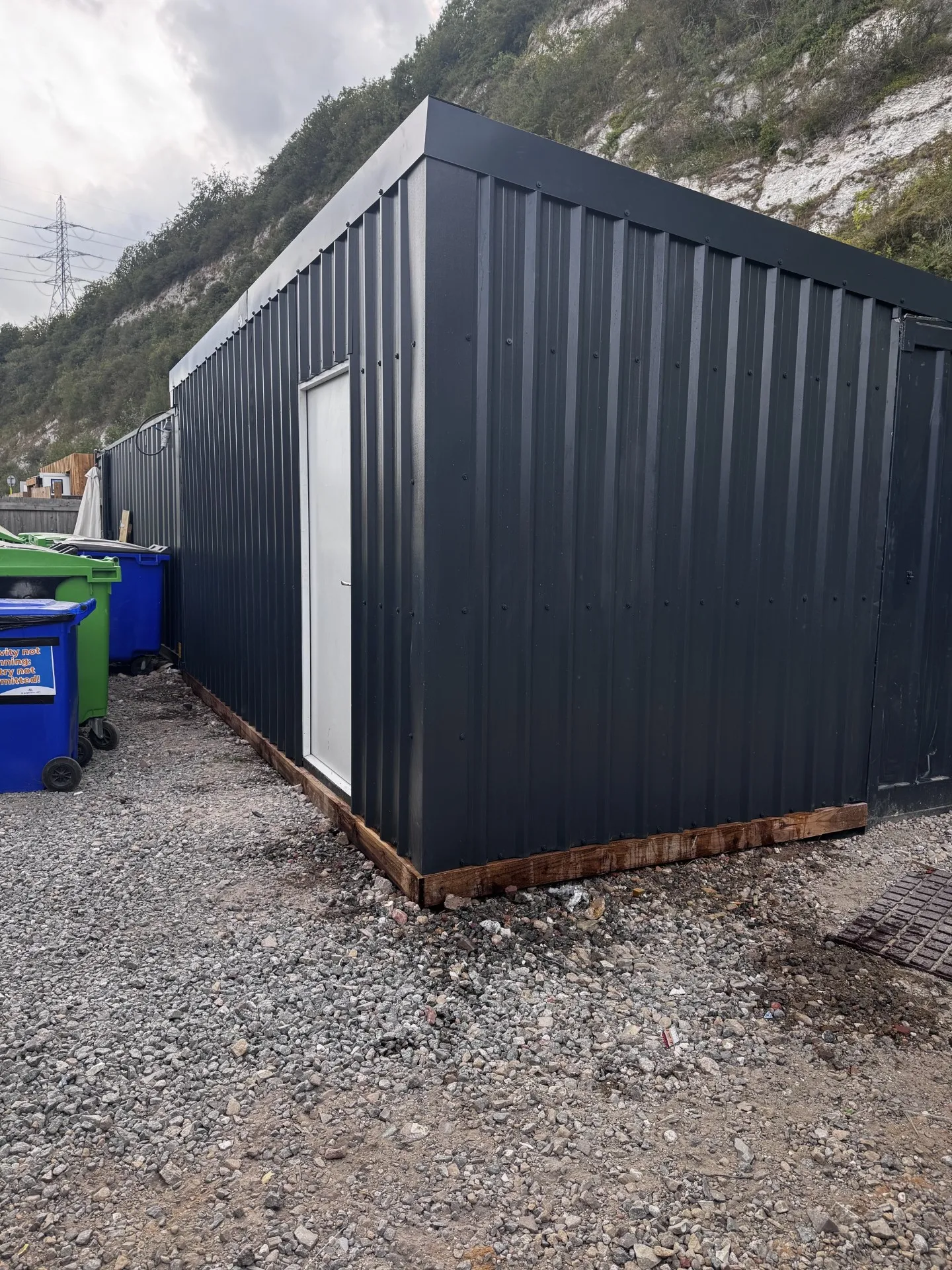 Dark grey airless-sprayed steel container with a white door, set on a gravel site with a chalk quarry backdrop